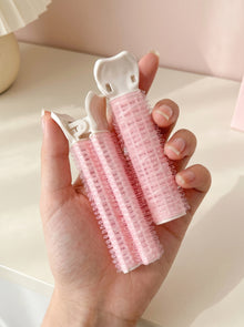 Dúo de productos rosas con pinza blanca para dar volumen al cabello, sostenidos en la mano sobre fondo claro.