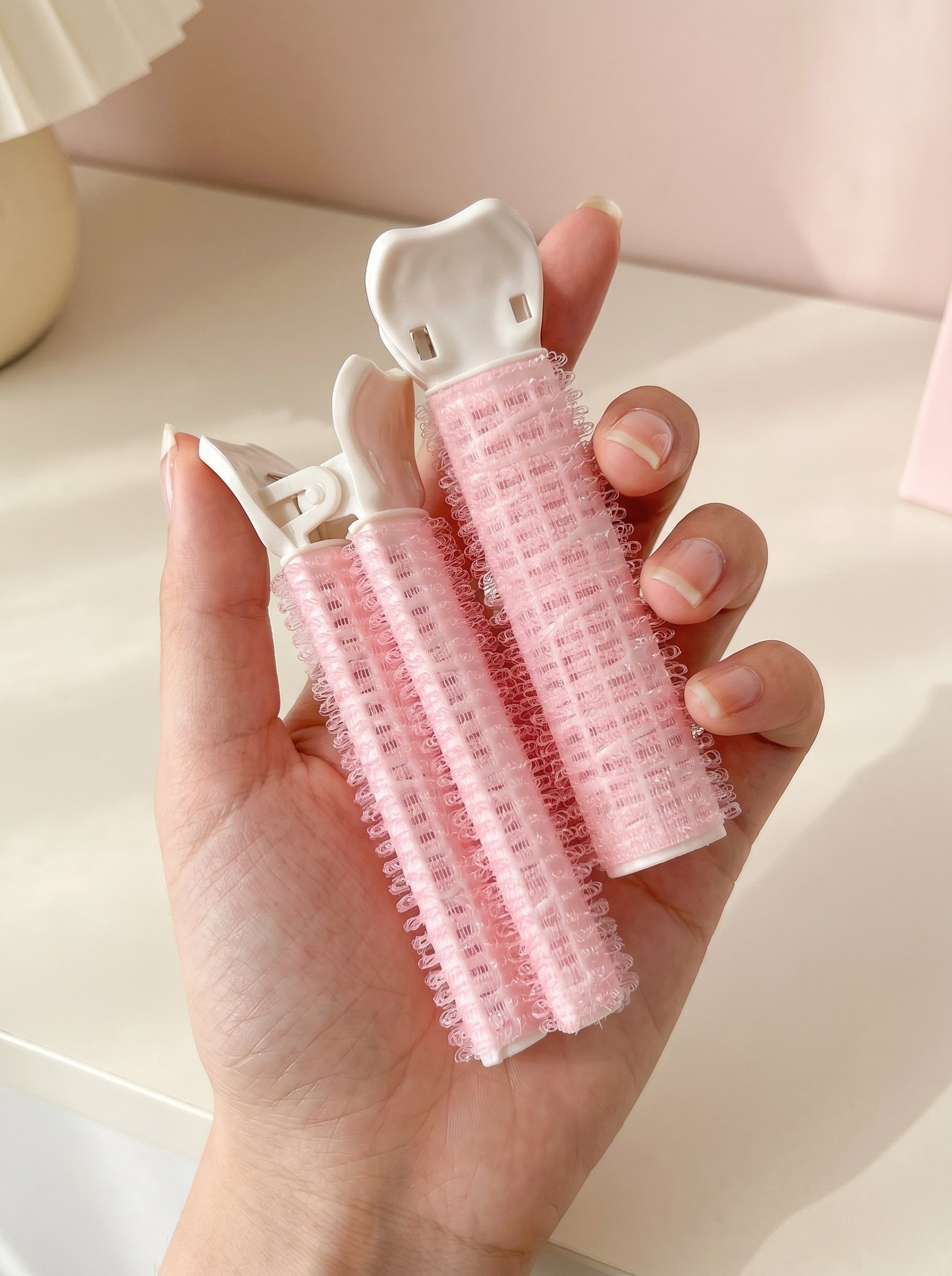 Dúo de productos rosas con pinza blanca para dar volumen al cabello, sostenidos en la mano sobre fondo claro.