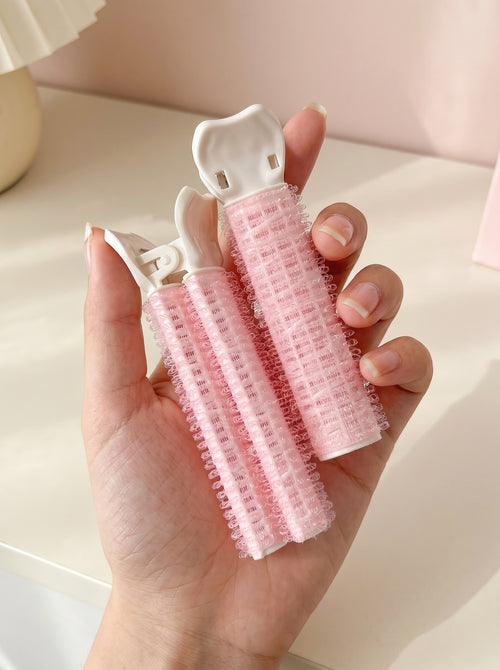 Dúo de productos rosas con pinza blanca para dar volumen al cabello, sostenidos en la mano sobre fondo claro.